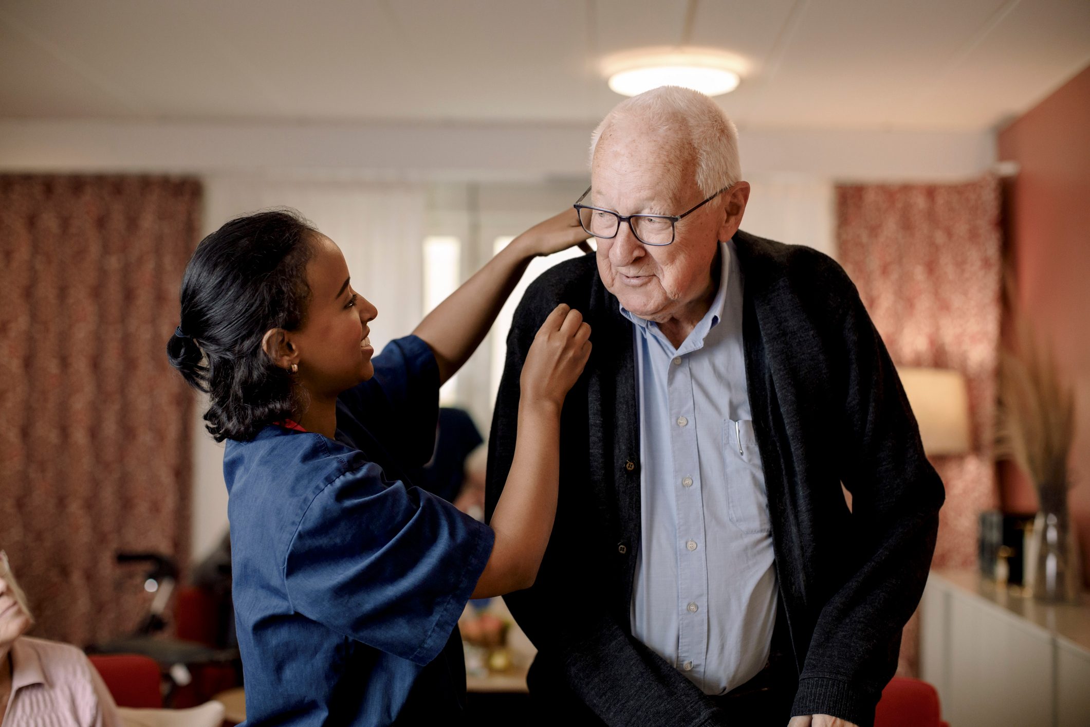 Senior man looking at female nurse helping him to wear sweater in retirement home