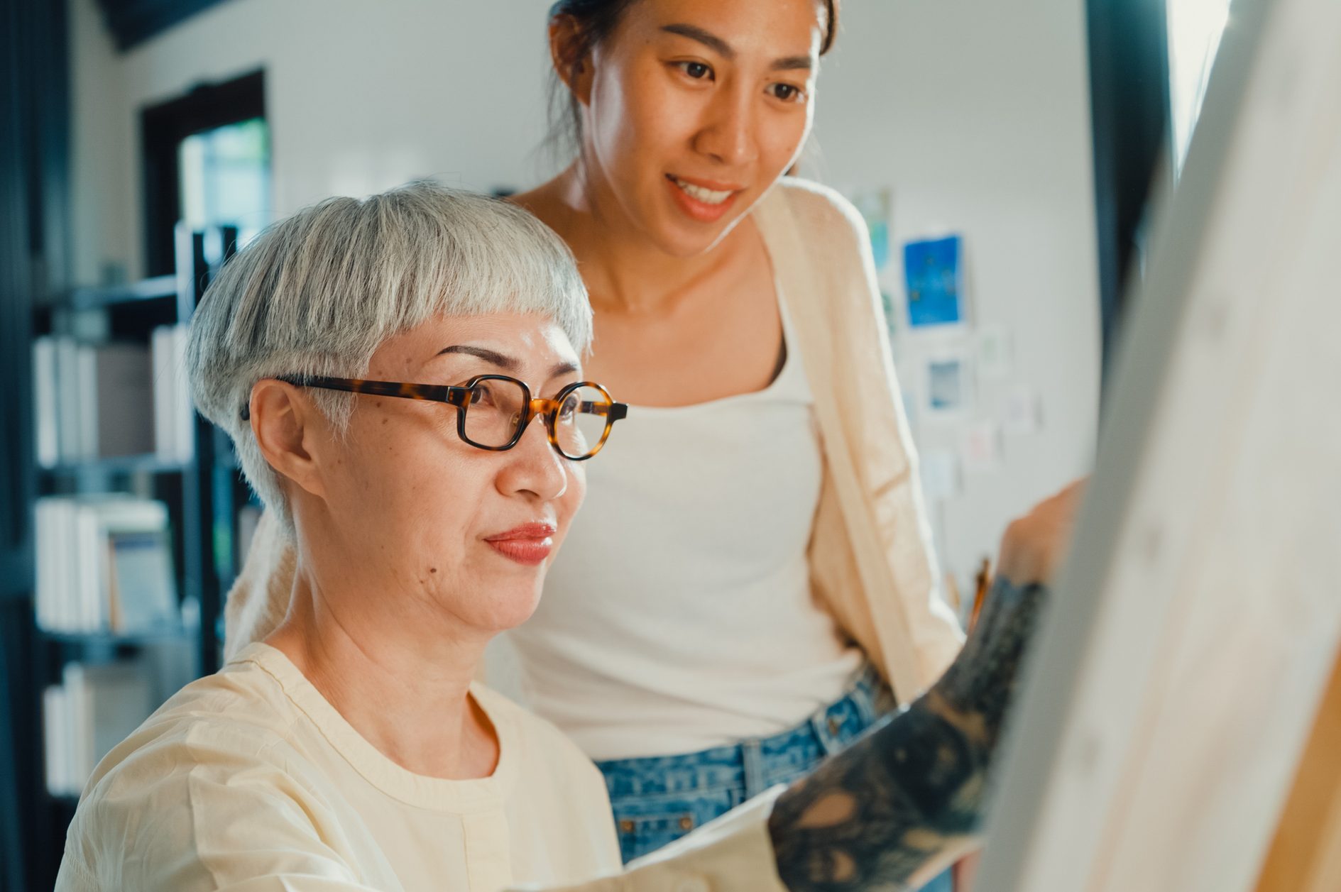 Close-up Happy Asian family daughter enjoy see older mother drawing painting colorful artwork on canvas spend time together in living room at home on weekend.