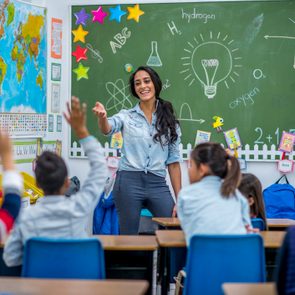 A teacher is leading a class of elementary school children. There are various posters on the wall, and drawings on the chalkboard. Students are putting up their hands to answer a question.