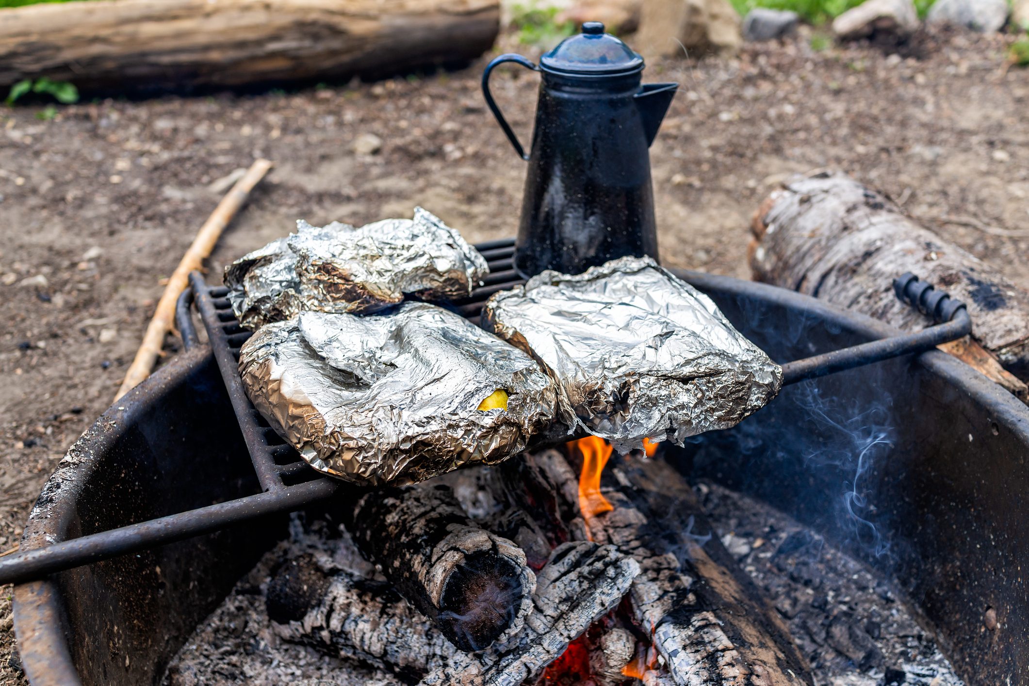 Blue tea water kettle and foil wrapped vegetables on grill in fire pit at campground with red flame fire burning in evening