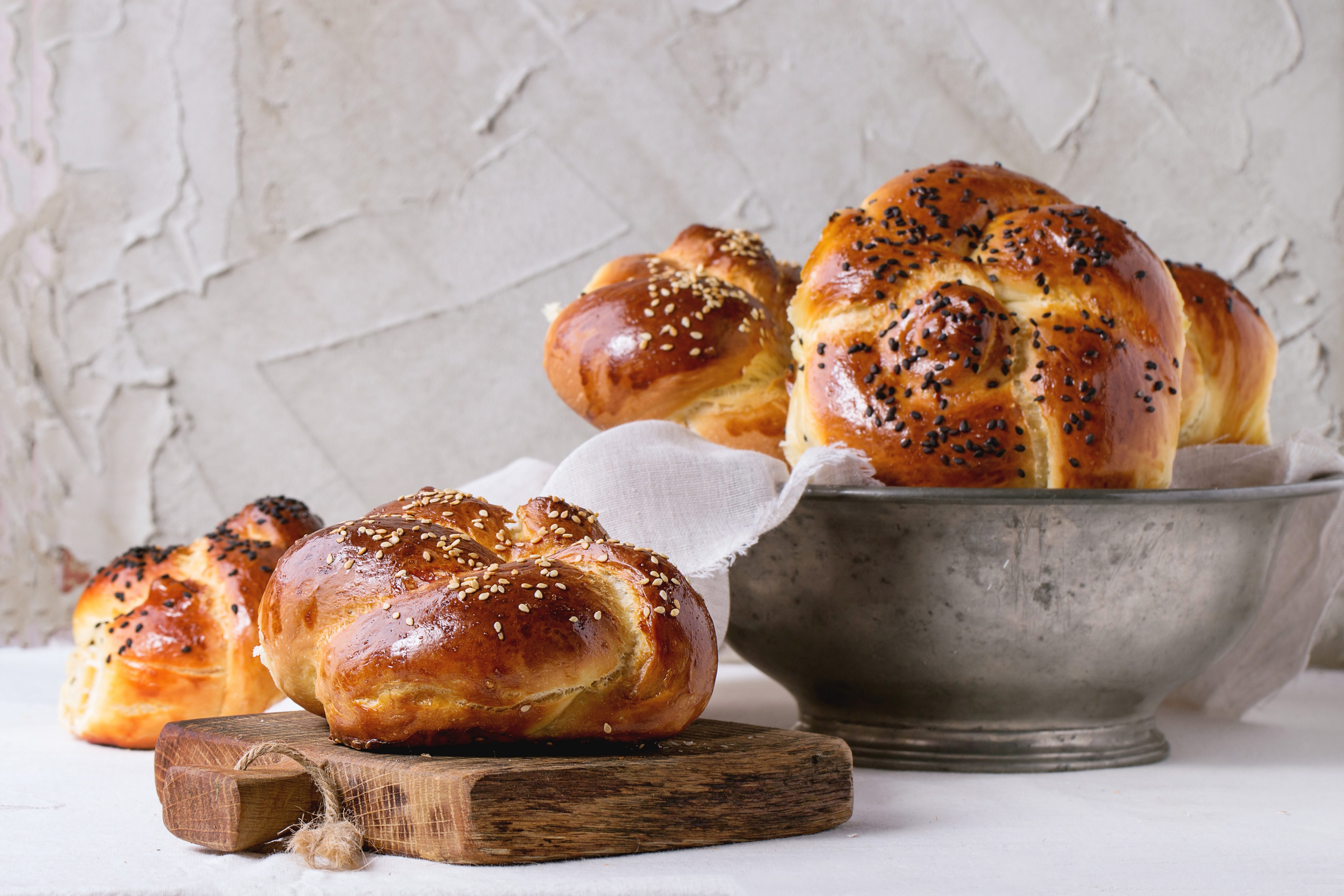 Heap of sweet round sabbath challah bread with white and black sesame seeds in vintage metal bowl and on small cutting board over white table with plastered wall at background.