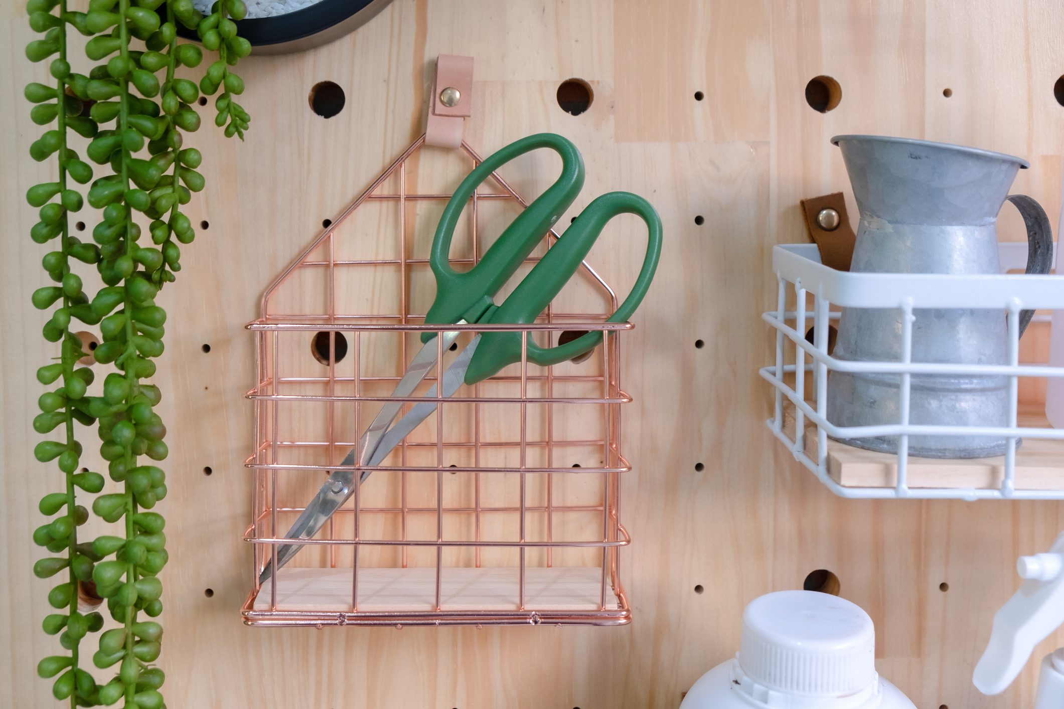 Green scissors in pink gold basket metal hanging on a Pine wood pegboard.