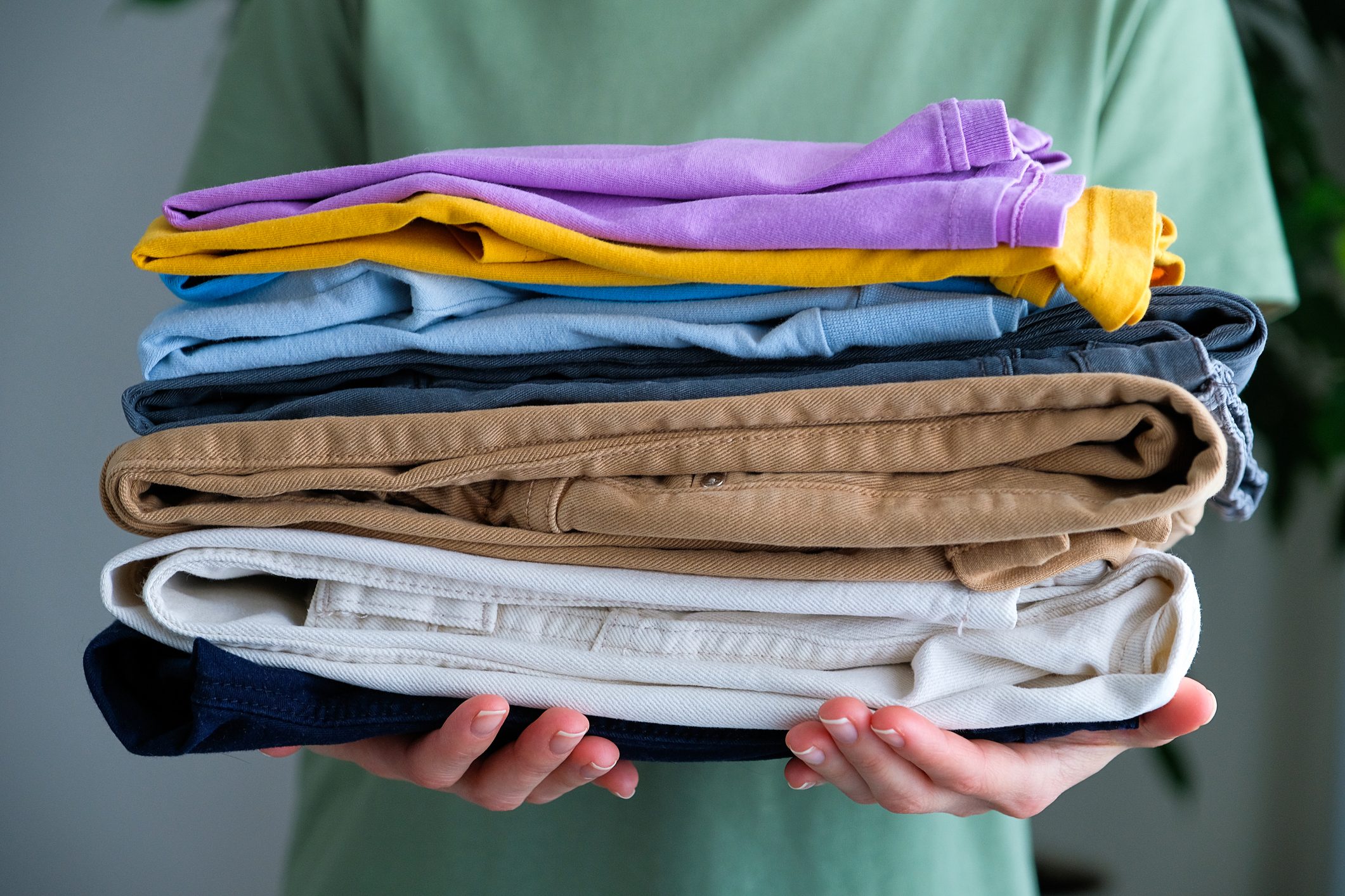 A female housekeeper or a girl holds a stack of things in her hands, at home or indoors. Clean folded cotton denim and bright colored T-shirts. The concept of housework, ironing, care and storage of things.