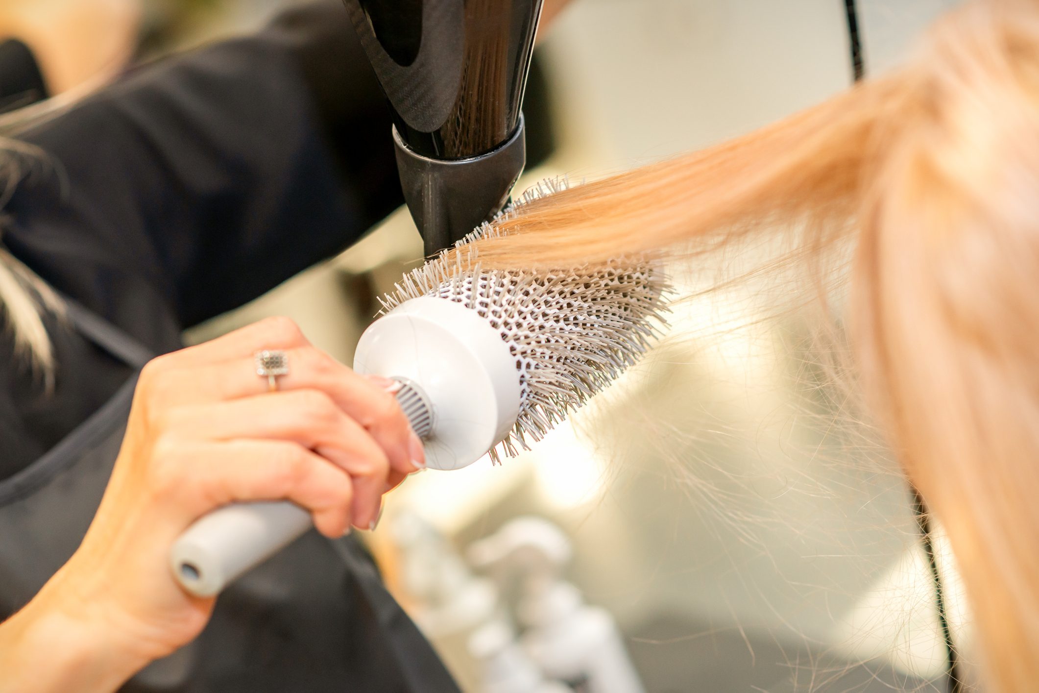 Drying straight blond hair with black hairdryer and white round brush in hairdresser salon, close up.
