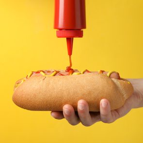 Person pouring ketchup on hot dog, on yellow background