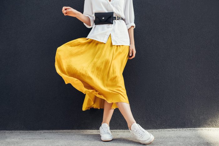 Horizontal cropped image of stylish slim woman in beautiful yellow skirt. Caucasian female fashion model standing over gray wall background outdoor with copy space.