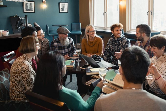Large group of people with a mixed age range sitting together around a table. They are enjoying a drink and having a book club meeting.