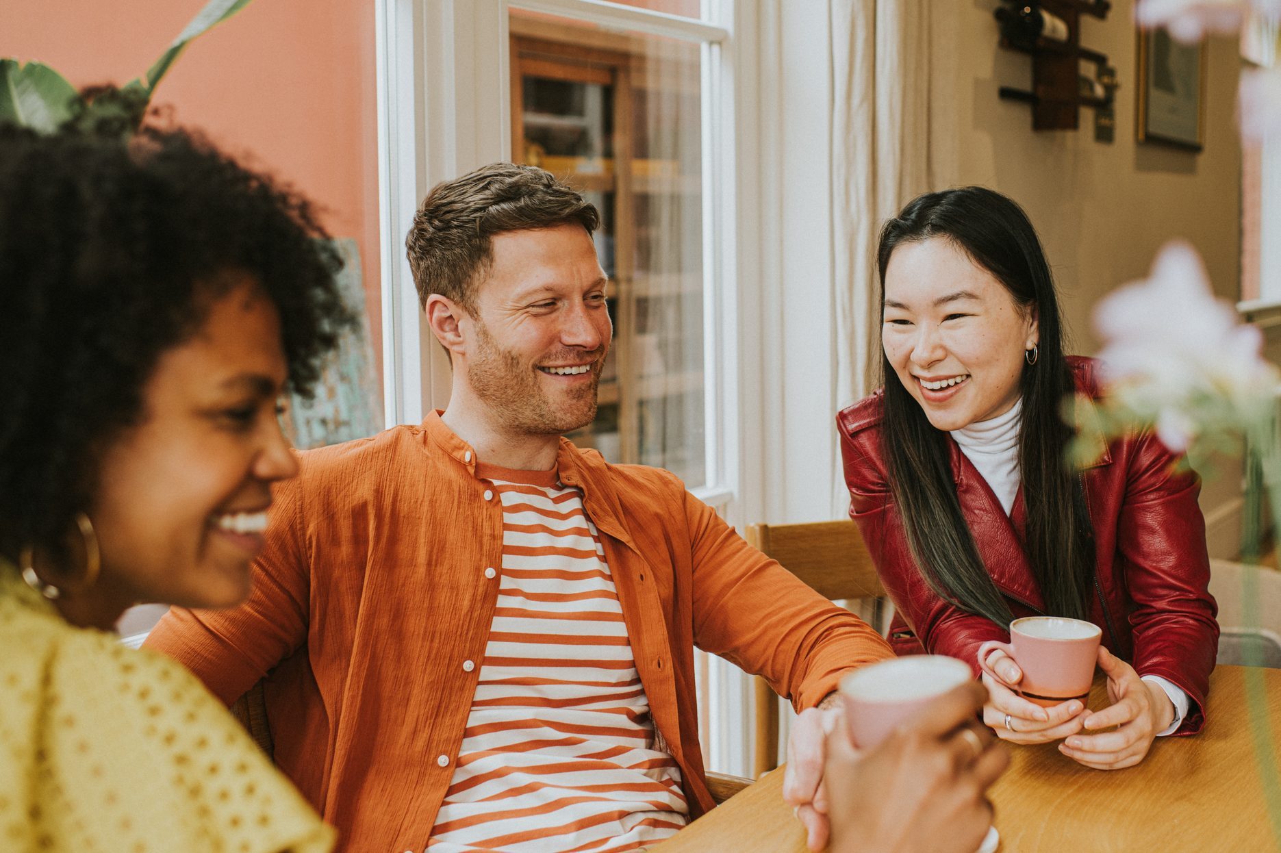 Three young people sit around a table and giggle as they have a lighthearted discussion and drink coffee / tea