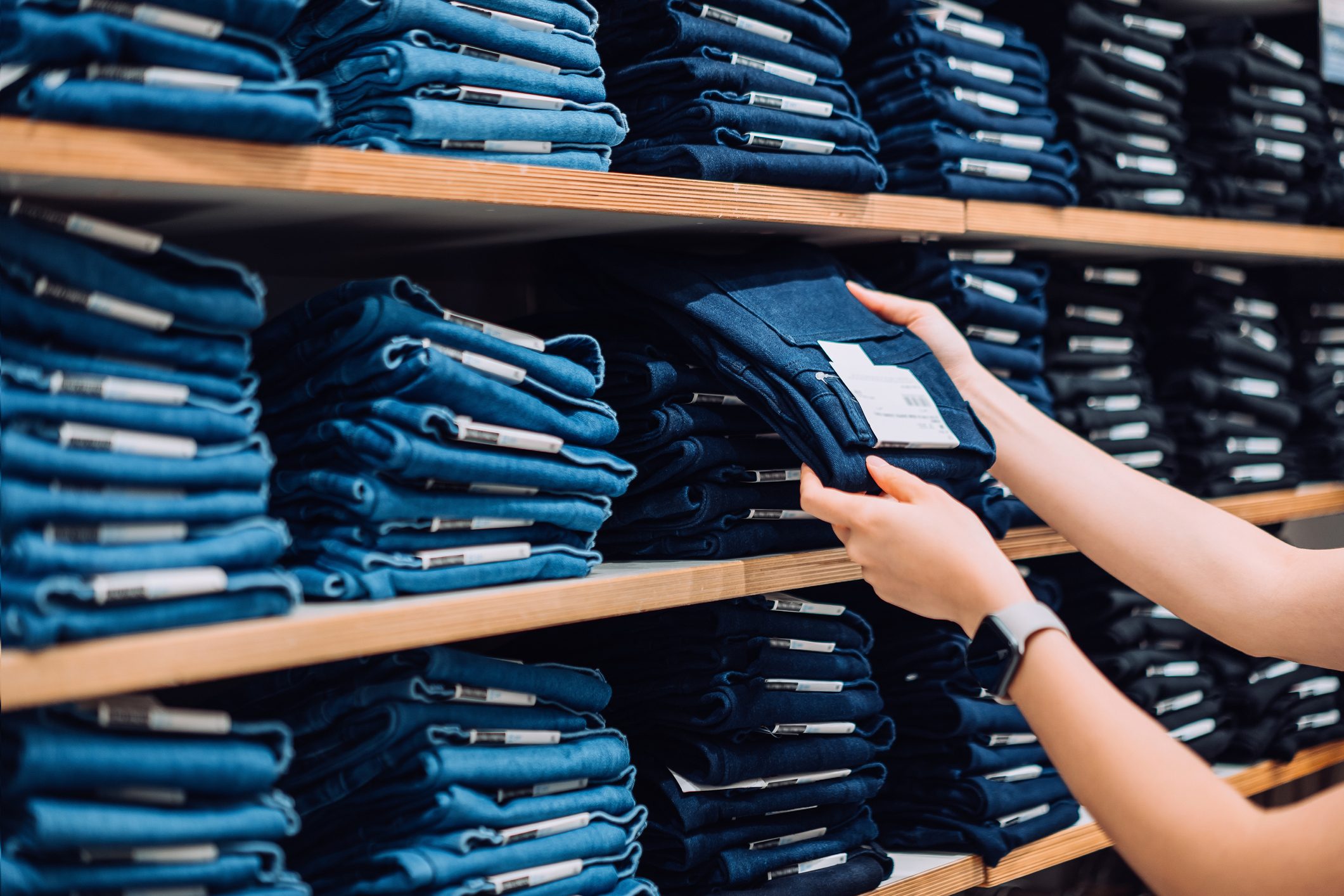 Cropped shot of woman's hand selecting a pair of trousers from the display shelf while shopping in a clothing store in the city