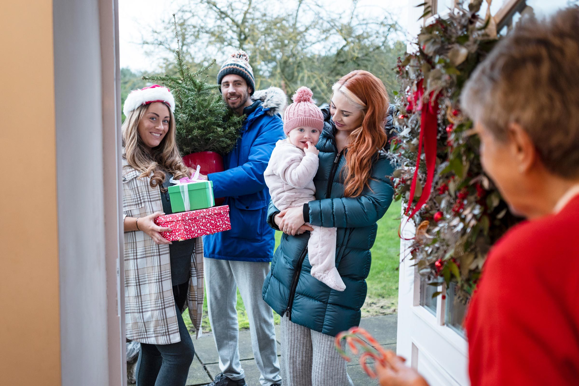 An over the shoulder, medium shot of a senior woman opening her front door to her three grandchildren and her great granddaughter at Christmas. They are standing outside holding gifts and a Christmas tree.