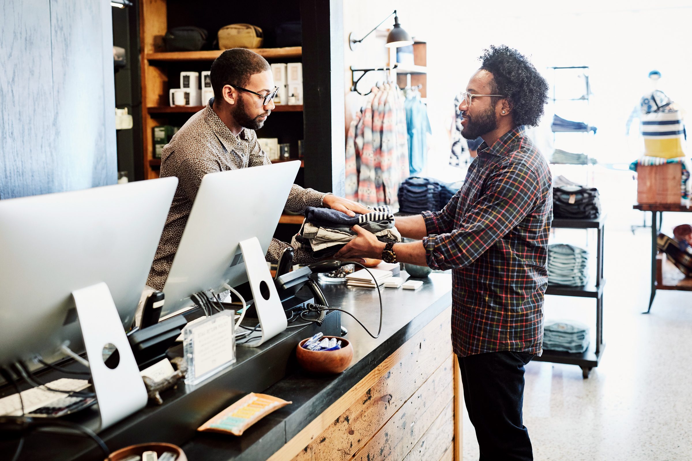 Man handing clothes to shopkeeper at register in mens clothing boutique