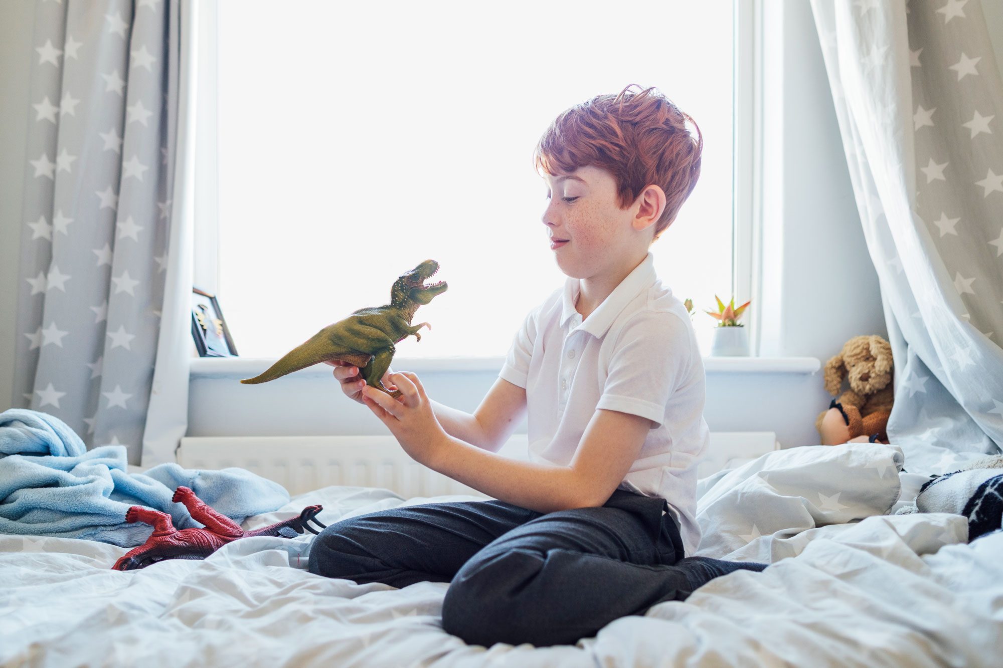 redhead boy playing with dinosaur toys on his bed in his bedroom at home, near a window