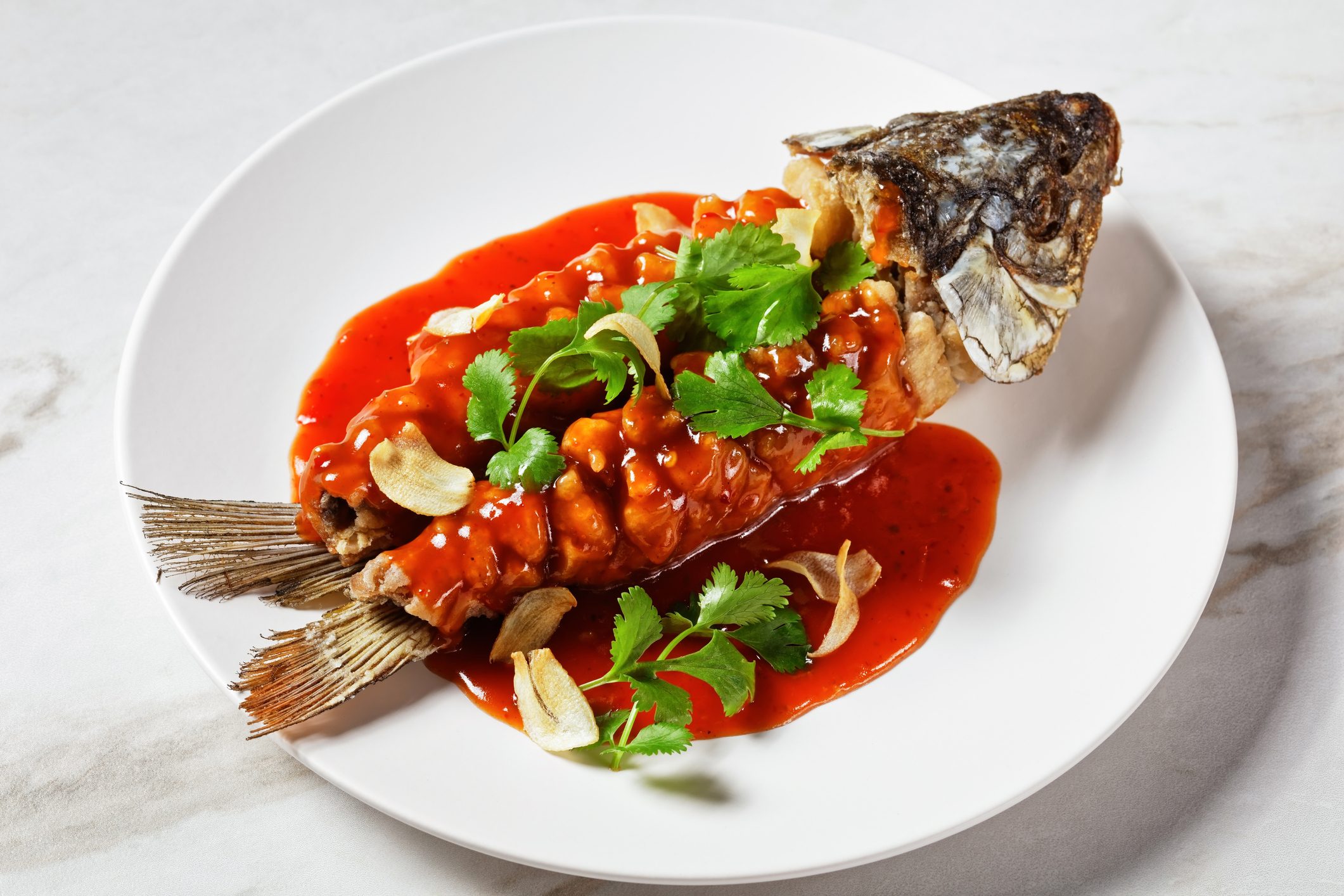 Chinese food: carp squirrel or songshu yu under sweet and sour sauce and fresh cilantro served on a white plate on a white marble stone background