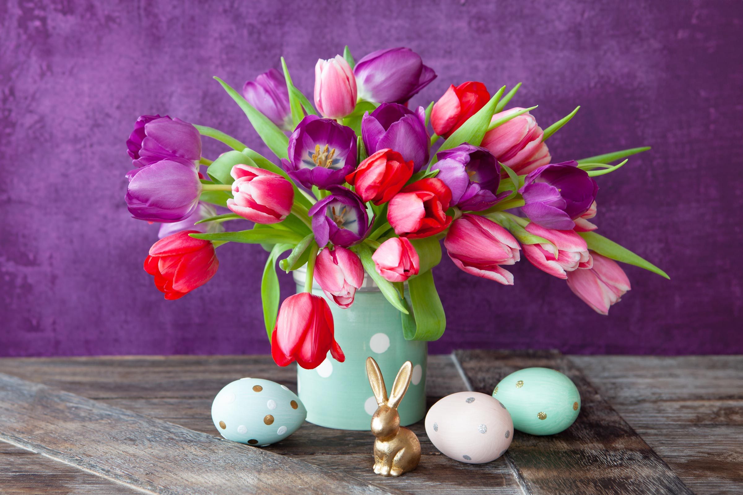 vase with red and purple tulips with pastel easter eggs and bunny direction on a wooden table with a purple background