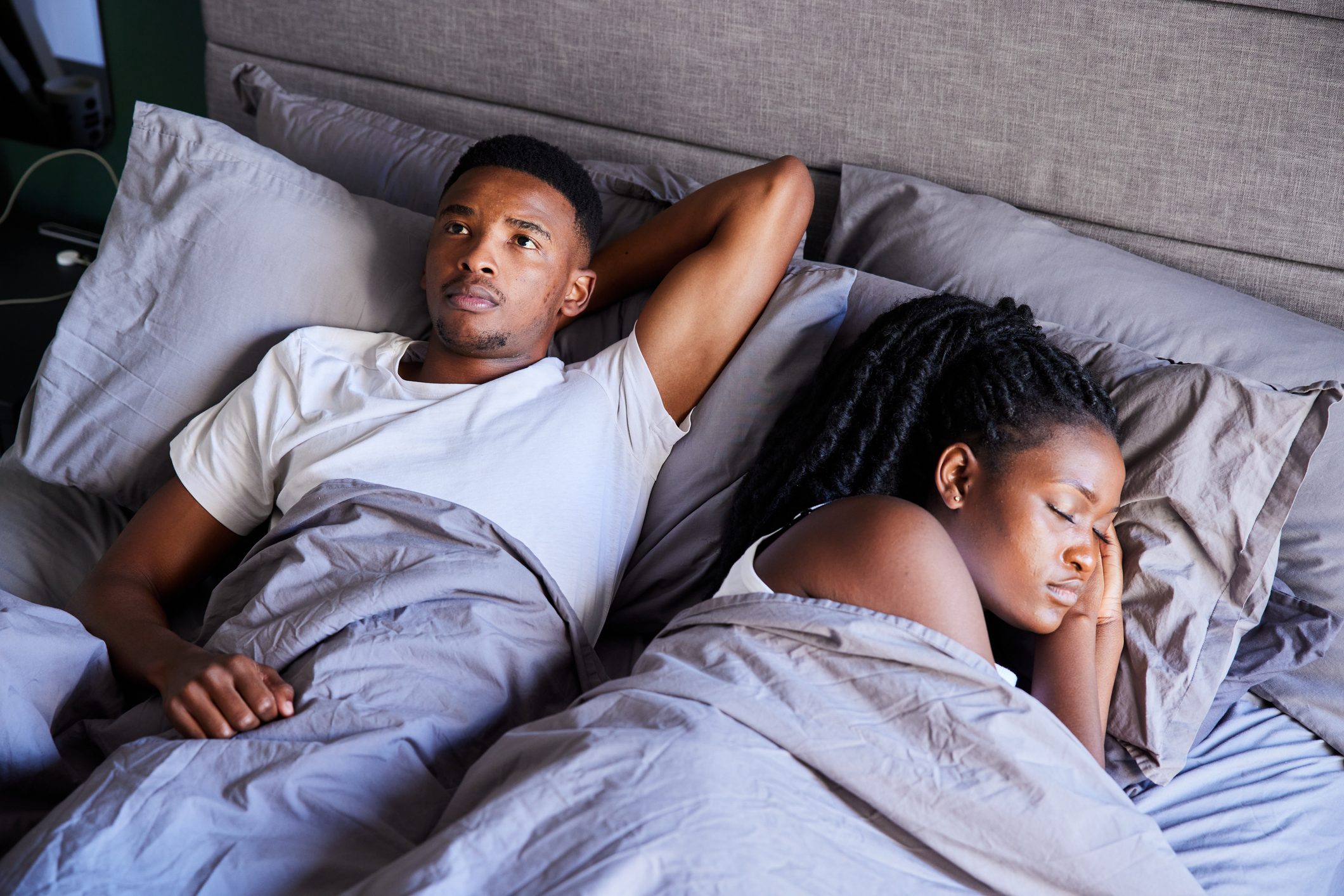 Young man looking unhappy while lying in bed with his sleeping wife