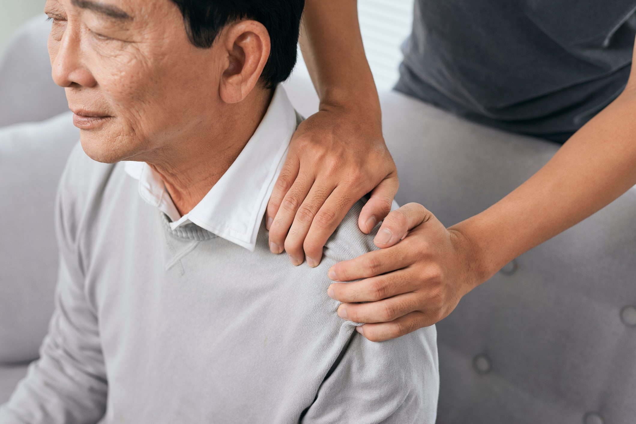 Asian old man shoulder pain, sitting on sofa , son massaging father shoulder.