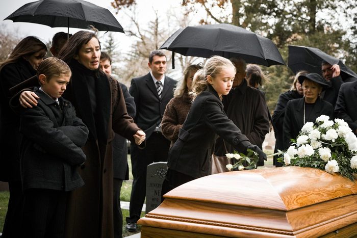 A funeral scene with mourners dressed in black, holding umbrellas as they stand in the rain. A young girl places white flowers on a wooden casket. Tombstones and bare trees are visible in the background, indicating a solemn, overcast atmosphere.