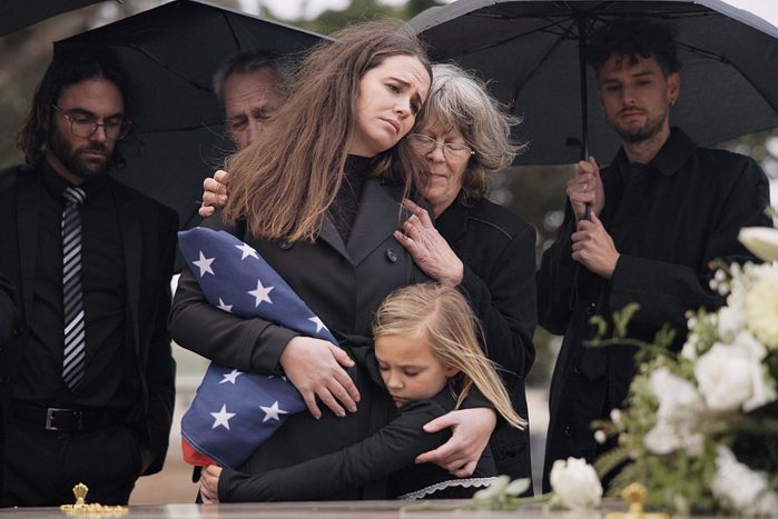 Funeral, family hug and sad people with grief support, goodbye service and mourning death at burial event. Kid child, mom embrace and group gathering together at coffin, casket and crying at ceremony