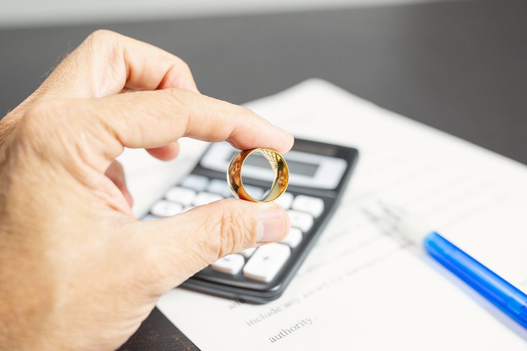 A hand is holding a gold ring in front of a calculator and some papers on a dark table. The focus is on the ring, with the calculator and documents slightly blurred in the background.