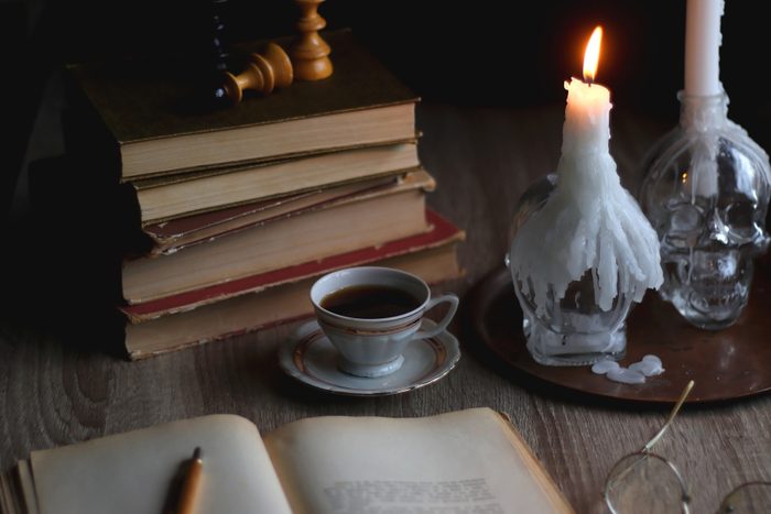 A candle burns in front of a pile of books. A cup of coffee on a saucer is between the two. An open book rests in the foreground and spectacles can be seen in the corner.