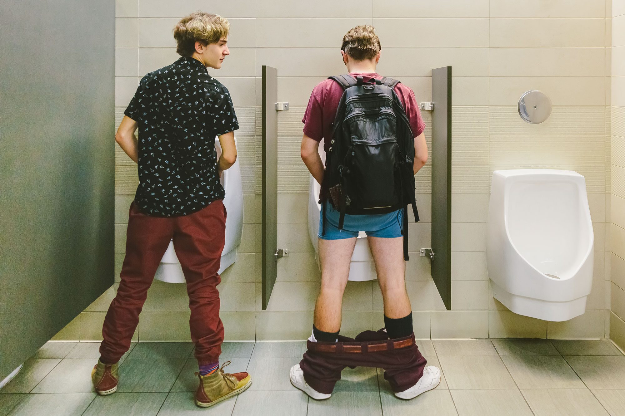 Man With Pants Pulled Down At Urinal While Another Man Looks At Him