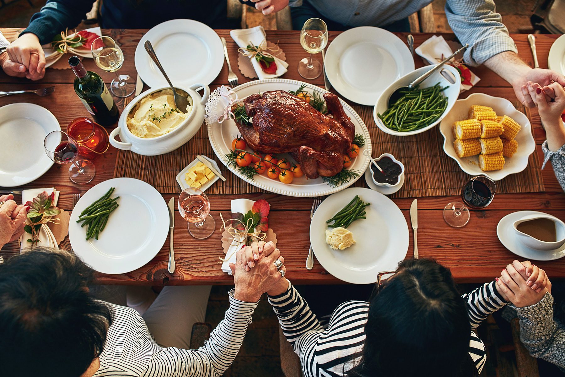 A group of people gathered around a wooden table for a meal, holding hands. The table is set with plates, cutlery, glasses, and bowls filled with green beans, mashed potatoes, corn, and a roasted turkey in the center.