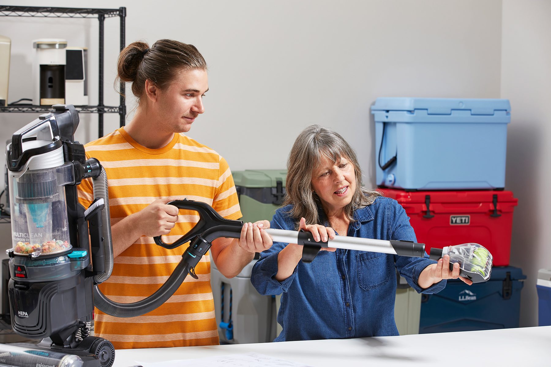 A young man in a striped shirt holds a vacuum cleaner handle while an older woman in a blue shirt adjusts it. They stand in a room with shelves and storage bins in the background.