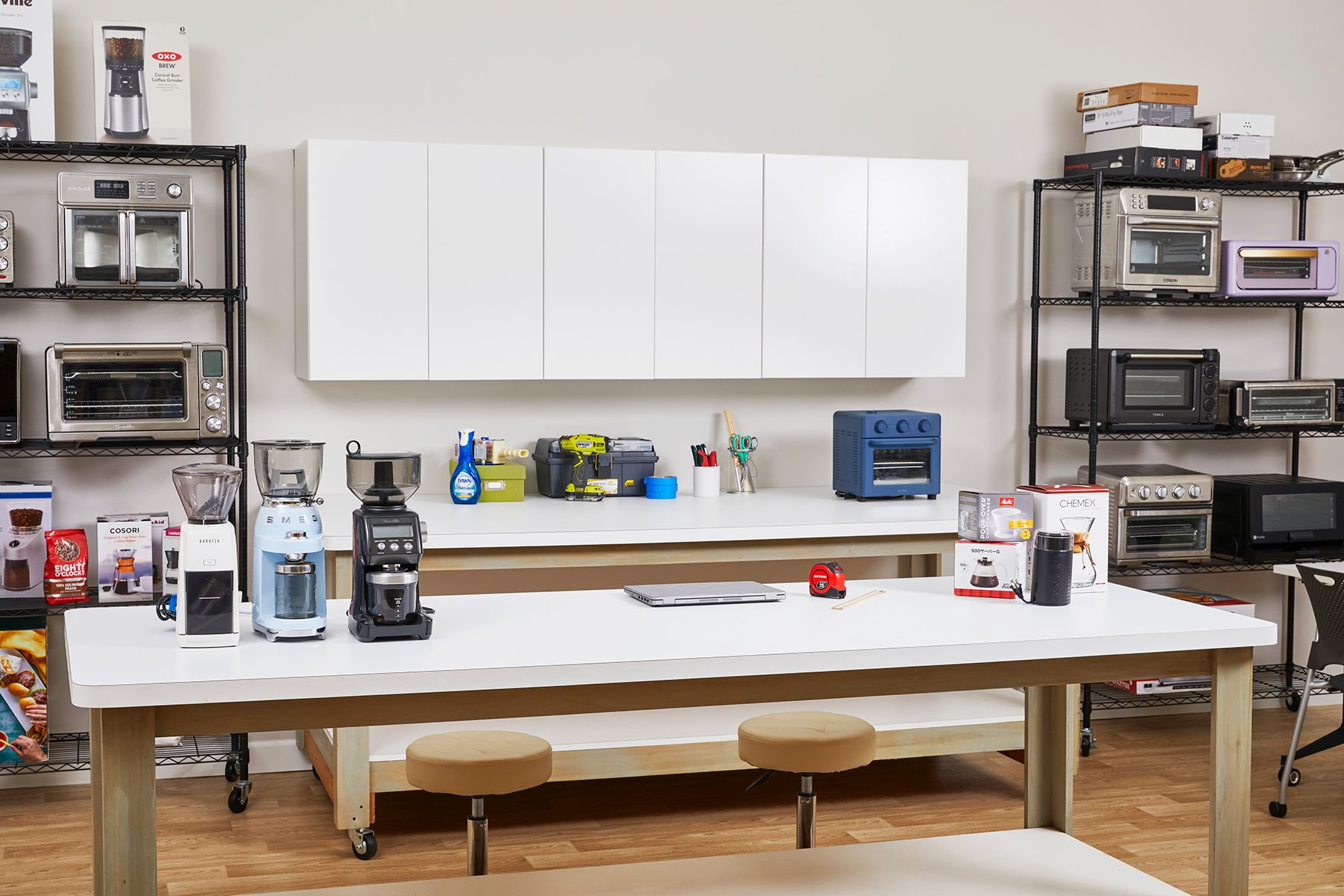 A kitchen showroom features two large tables with various kitchen appliances and gadgets on display, including coffee makers and blenders. Shelves on the walls hold items like toasters and microwaves, while white cabinets are mounted above the main counter.