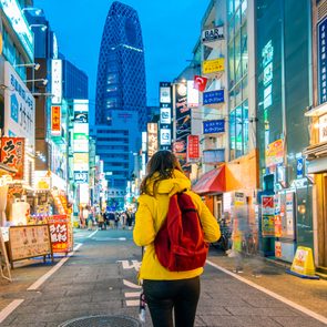 Person in yellow jacket with red backpack strolls down a lively street at night with a skyscraper in the distance