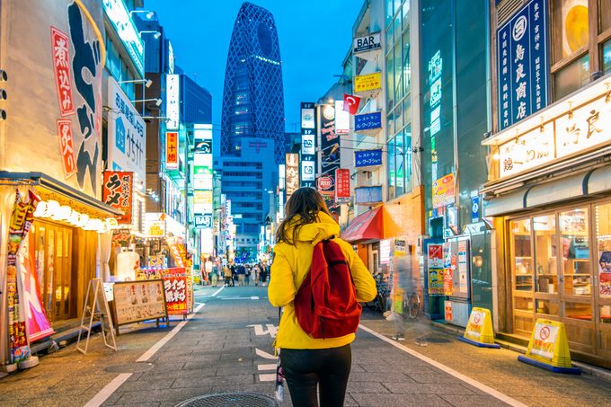 Person in yellow jacket with red backpack strolls down a lively street at night with a skyscraper in the distance