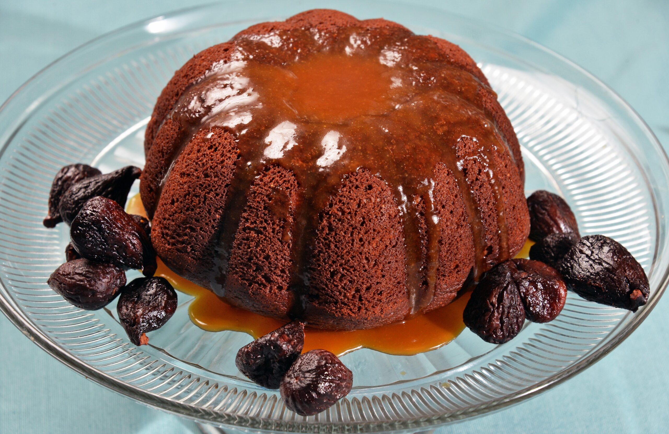 A brown cake draped in sauce sits on a clear cake stand accompanied by dried figs