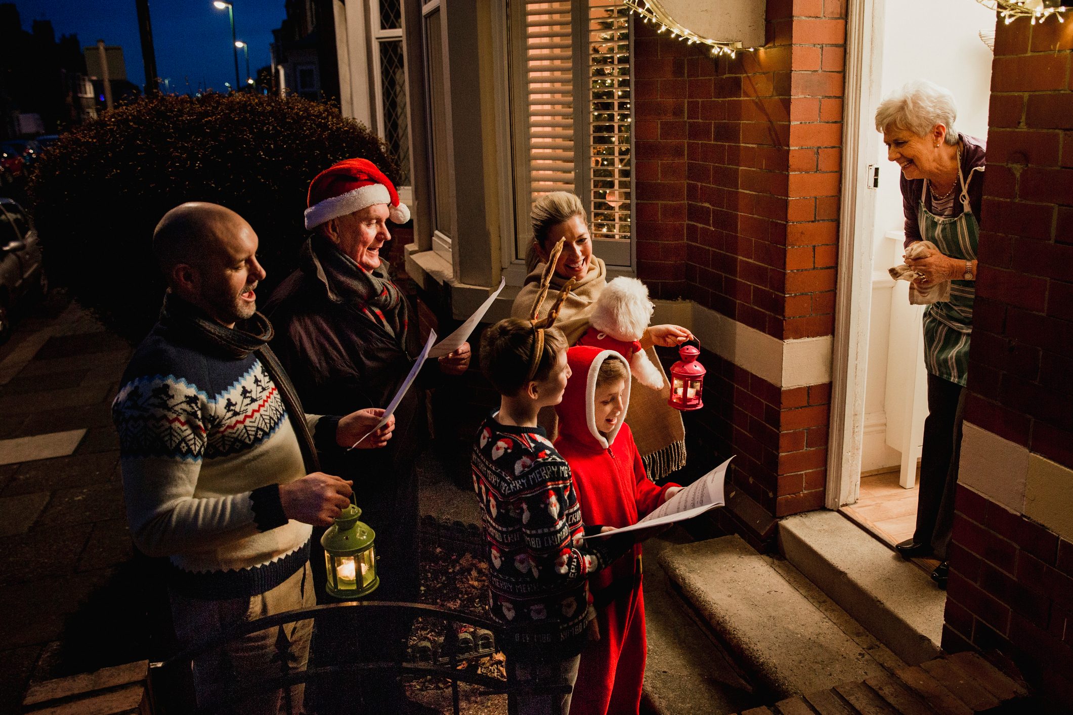 A family dressed in festive attire singing Christmas carols to an older woman in her doorway