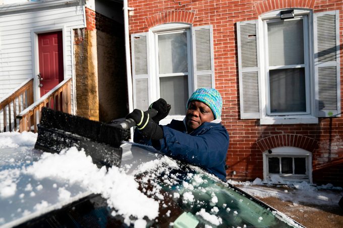 Woman in a turquoise hat and navy coat scrapes snow off the roof of her silver car with a brick house in the background