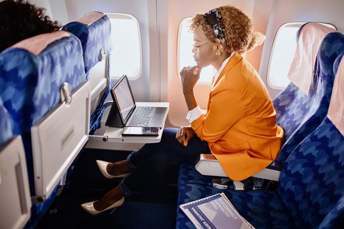 Woman in a window seat of an airplane working on her laptop during a flight. The middle seat next to her is free and she's placed some items on the seat.