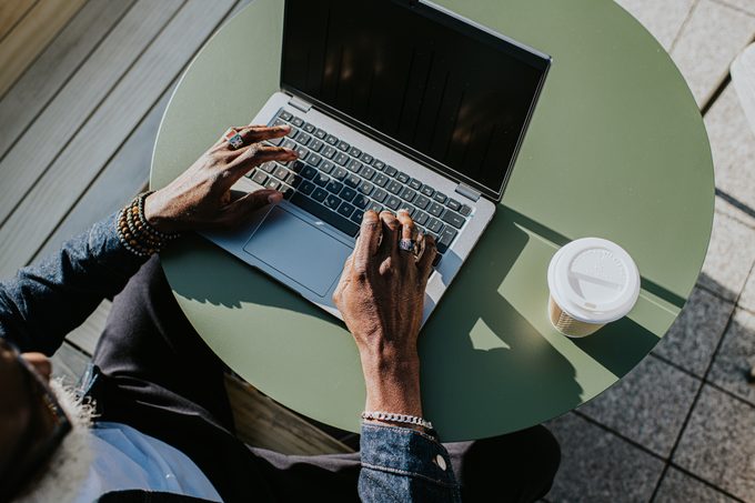Close-up of hands typing on a laptop computer on a green outdoor table with a cup of coffee to the right