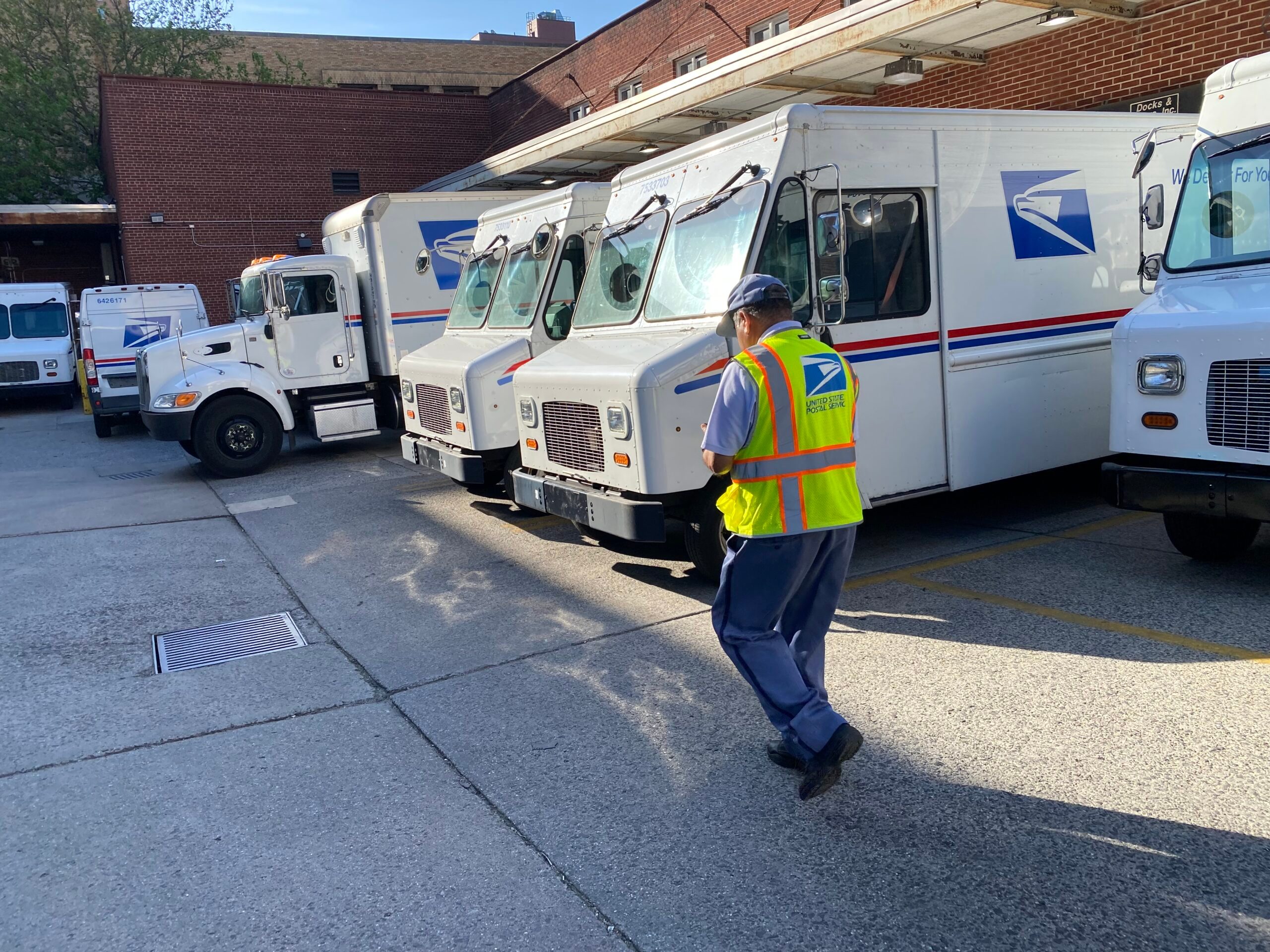 Mail delivery person in safety vest walking towards USPS delivery trucks