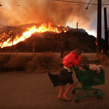 Woman pushes a grocery cart full of her belongings on a road with a hill on fire in the background
