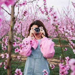 Young Woman Taking Photos Of Blossoming Fruit Trees With A Retro Camera In Spring