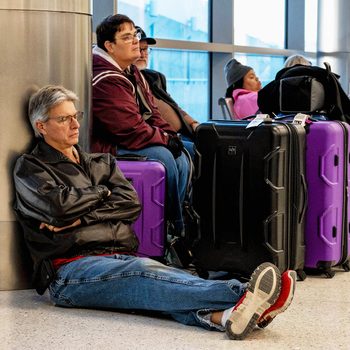 Man sits on the floor of an airport with eyes closed and arms crossed next to purple and black luggage