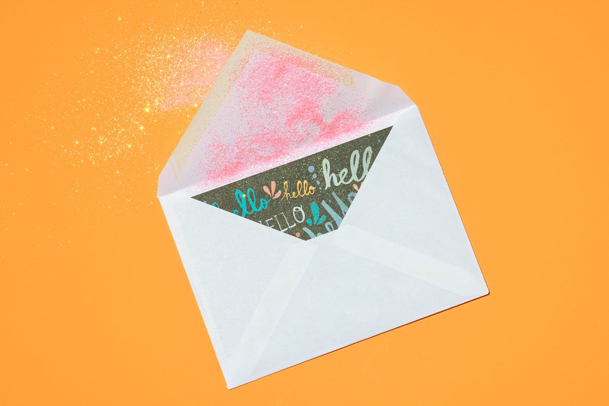 Overhead shot of a little sparkle card/invitation envelope with some glitter spilling out; white colored envelope; orange surface;