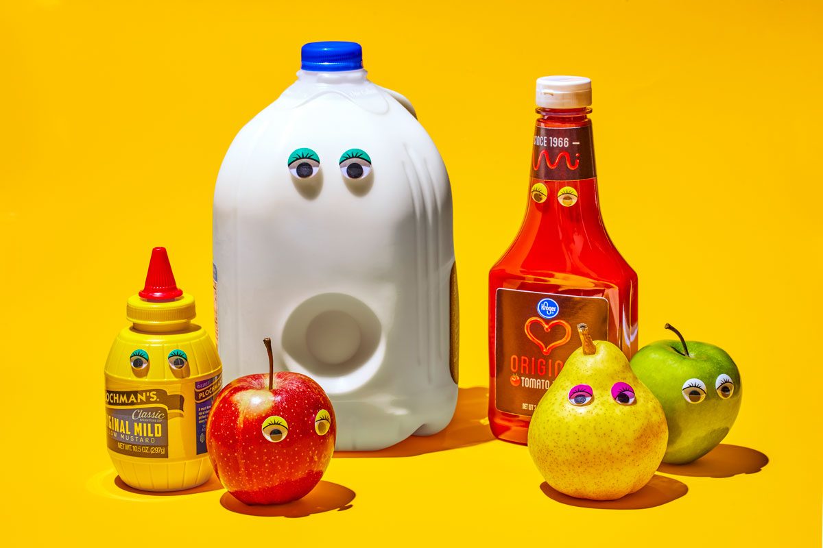 horizontal shot; A playful still life featuring common household items with googly eyes; A large milk carton, a ketchup bottle, a mustard bottle, a red apple, and a green pear are arranged on a vibrant yellow background; The addition of googly eyes to these items creates a whimsical and humorous composition;