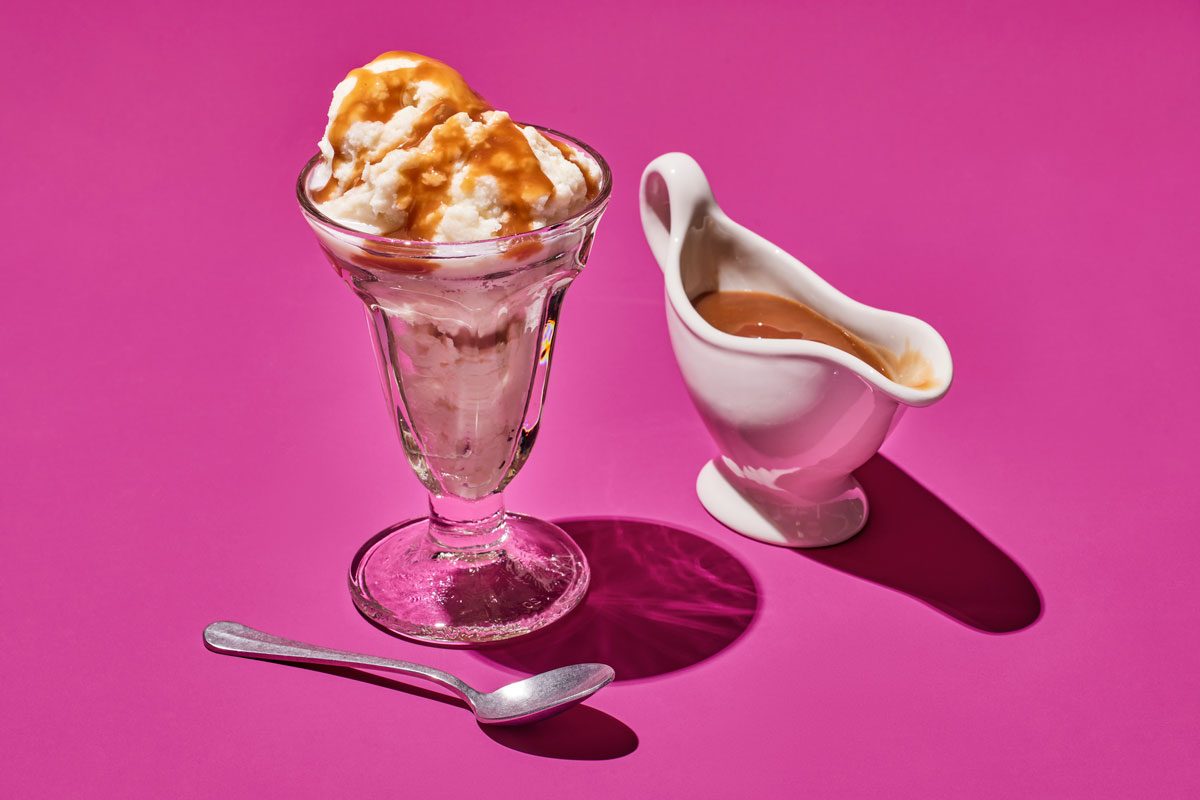 3/4th shot; A sundae glass is filled with creamy vanilla ice cream, drizzled with a rich, caramel sauce; The ice cream is topped with a dollop of whipped cream, if desired, A small, white gravy boat filled with additional caramel sauce sits beside the sundae; A silver spoon rests on a bright pink background;