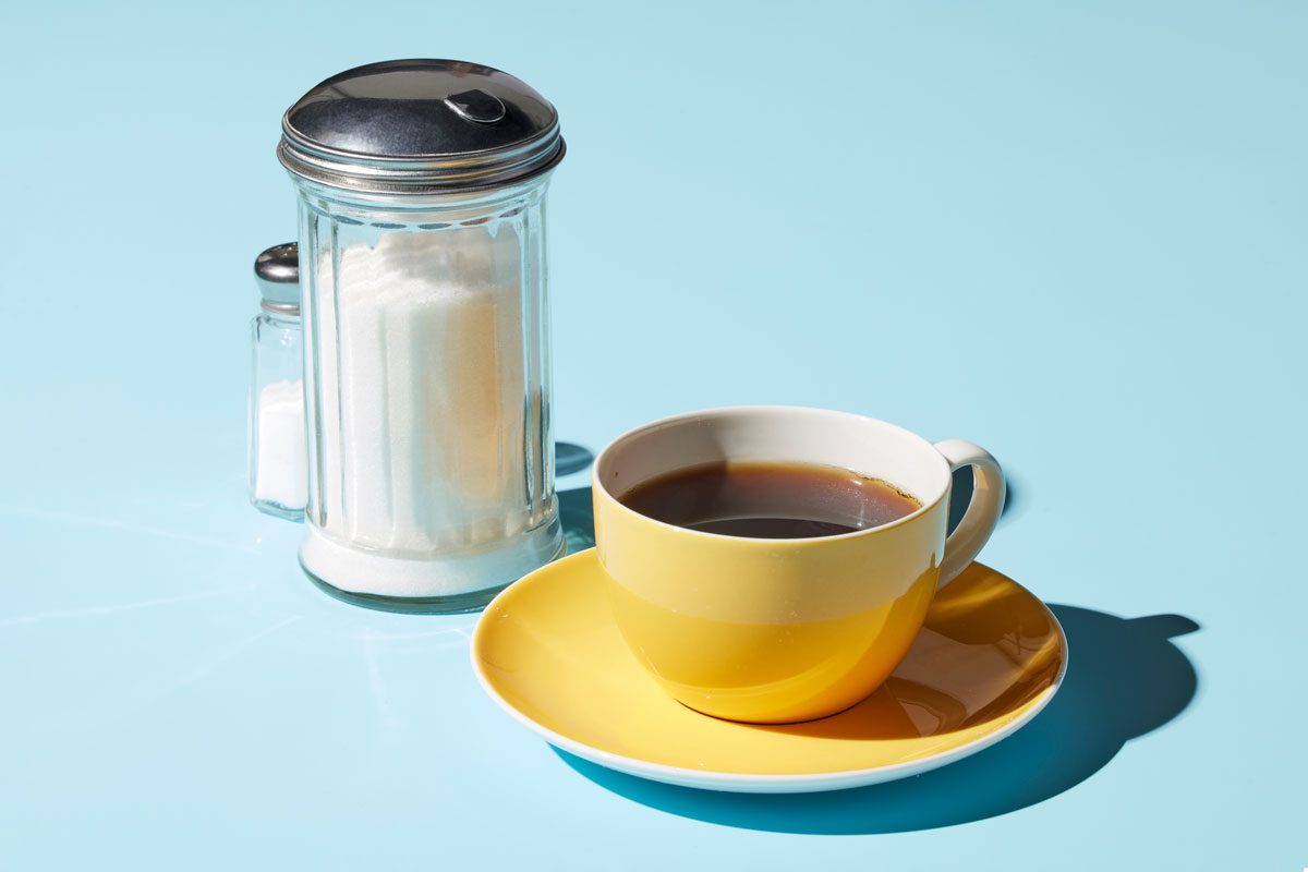 Table view shot of a cup of black coffee on a saucer; nearby a sugar shaker is positioned with a salt shaker peering out from behind; blue surface;