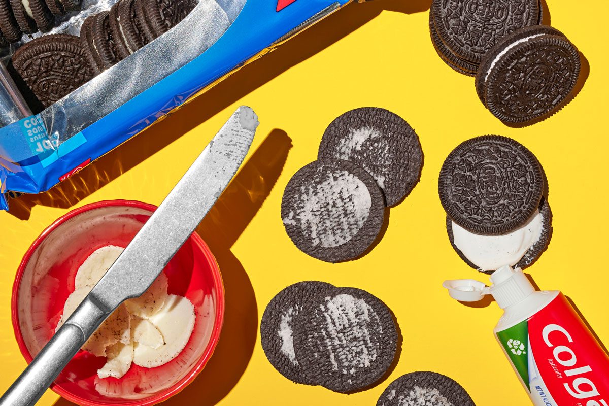 Overhead shot of unscrewed Oreos with discarded frosting and a butter knife nearby; a toothpaste tube is placed in place of the frosting; yellow surface