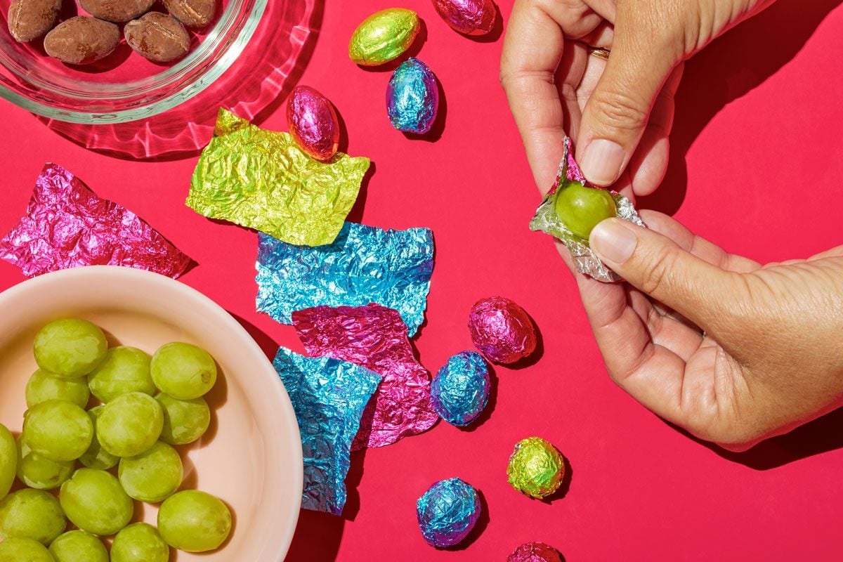 Overhead shot of foil wrapped chocolate eggs; some unwrapped foils; bowl for discarded chocolates; some grapes; grape about to be wrapped; and some already wrapped grapes; red surface;