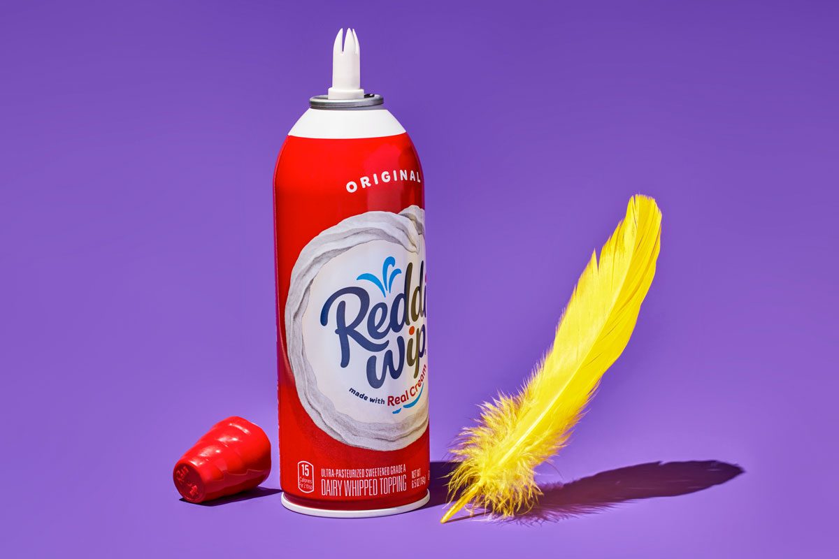 horizontal shot; A can of Reddi-wip Original Whipped Cream is the focal point of the image; The can is red with white text and features a playful design; A yellow feather rests beside the can; red cap of the can is placed on the left side of the can; The background is a vibrant purple;