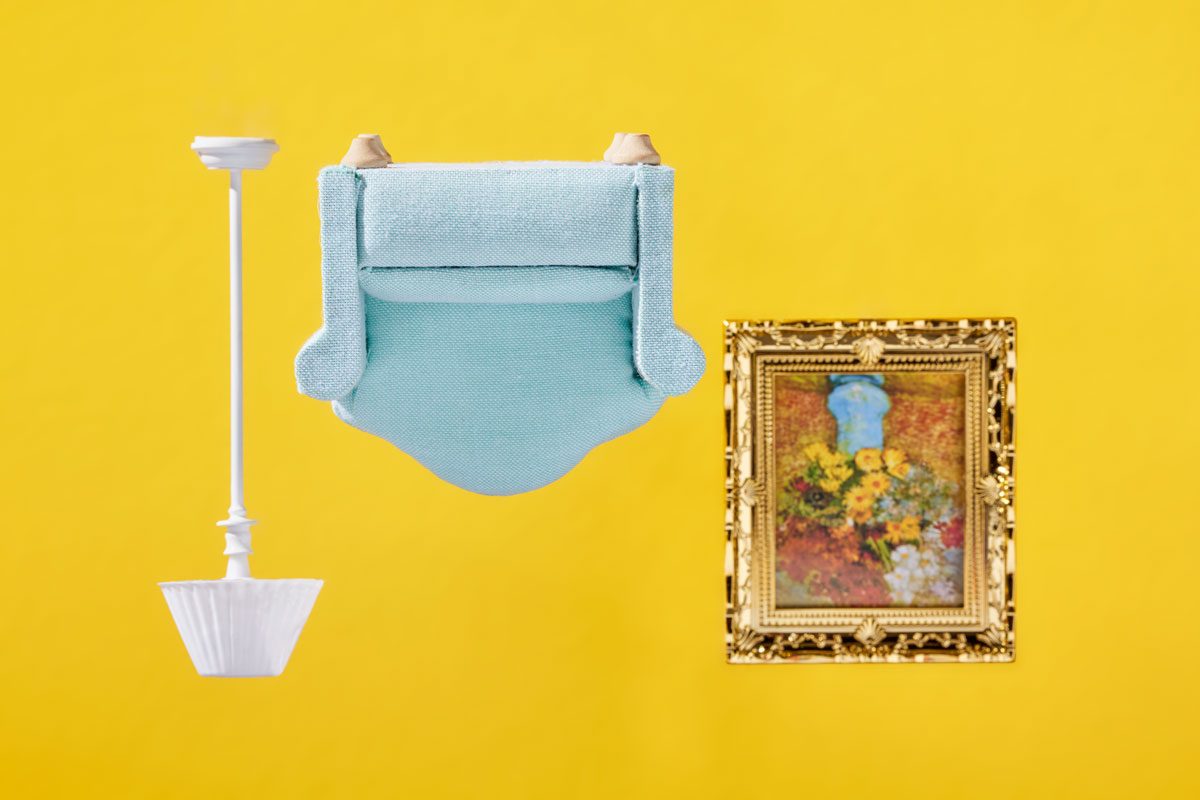 Front shot of an upside down house scene; featuring a dollhouse-sized armchair; lamp; and painting all floating in an inverted position; yellow background;