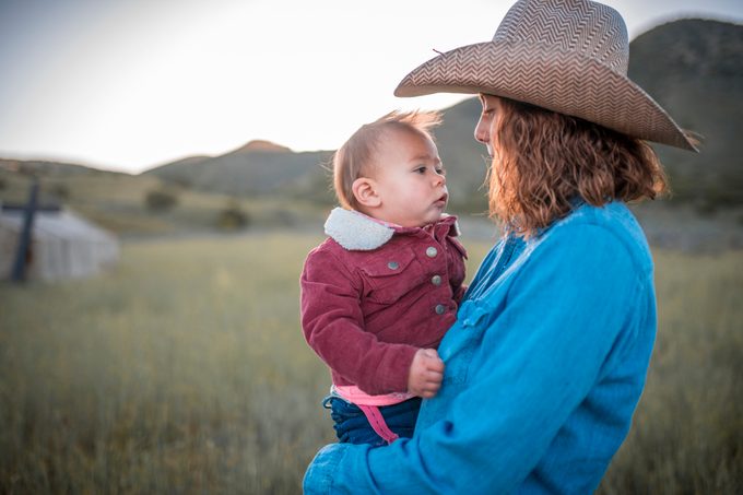 Woman in denim shirt and cowboy hat holds a baby in a red sheepskin jacket with mountains in the background