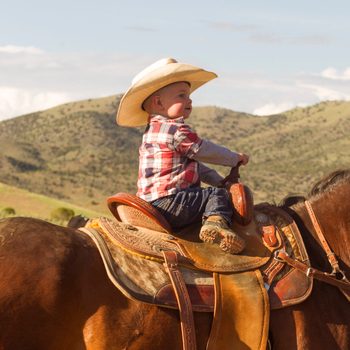 Baby in a tan cowboy hat and flannel shirt riding a horse with mountains in the background