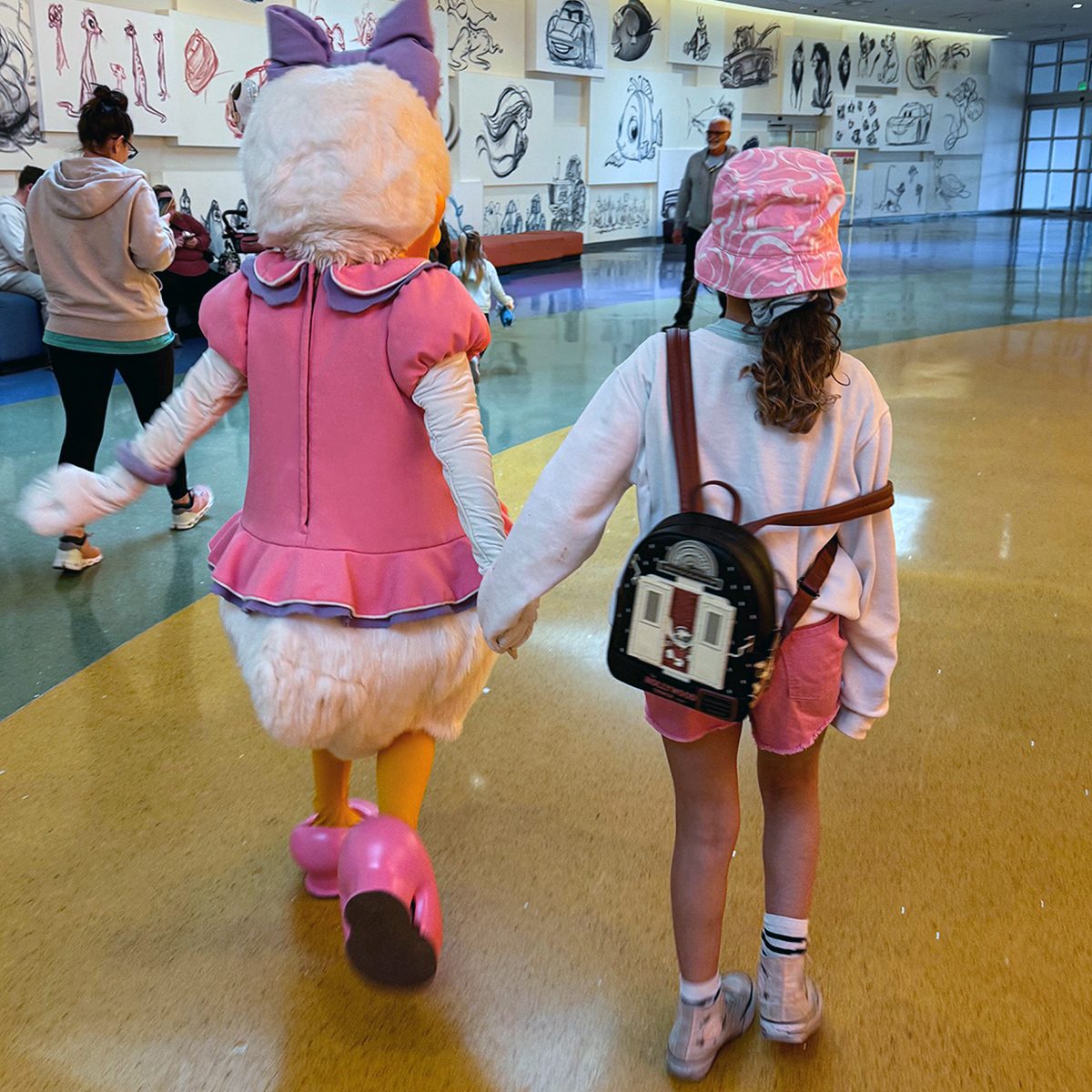 A person dressed in a pink character costume holds hands with a child wearing a pink hat, white shirt, pink shorts, and a backpack. They walk on a shiny floor with sketches on the walls around them.